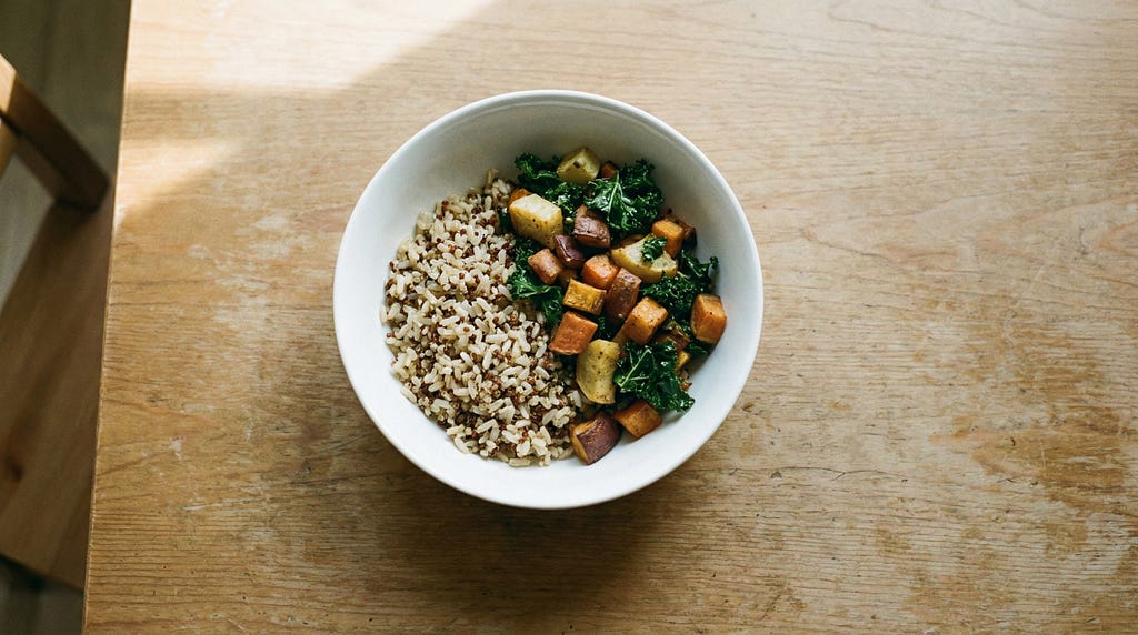 A simple bowl of grains and vegetables on a bare wooden table in soft natural light